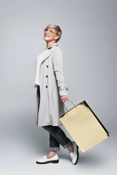 Smiling woman in stylish trench coat posing with shiny shopping bag on grey — Stock Photo