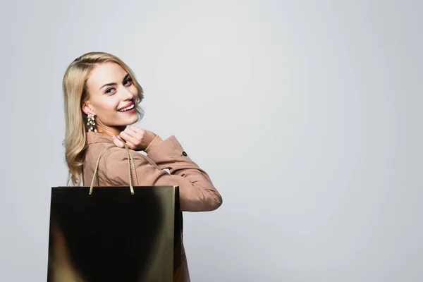 Joyful blonde woman with shopping bag smiling at camera isolated on grey — Stock Photo