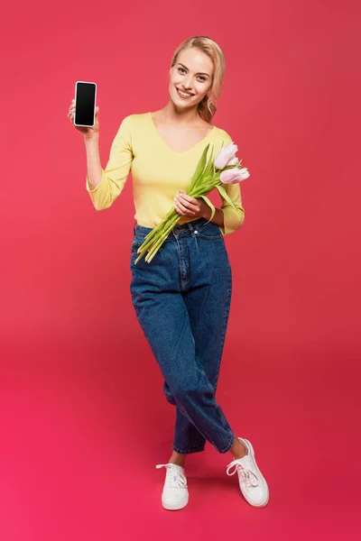 Blonde woman in yellow jumper and jeans holding smartphone with blank screen and tulips on pink — Foto stock