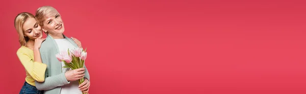 Улыбающаяся женщина, опираясь на радостную мать, держа тюльпаны изолированы на розовый, баннер — стоковое фото