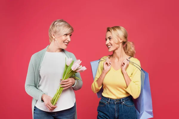 Cheerful mother and daughter with tulips and shopping bags smiling at each other isolated on pink - foto de stock
