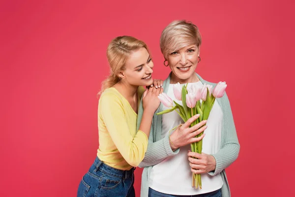 Smiling woman leaning on shoulder of joyful mother holding tulips isolated on pink — Stock Photo