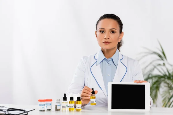 Médico afroamericano sosteniendo tableta digital con pantalla en blanco cerca de botellas con cannabis medicinal y cbd - foto de stock