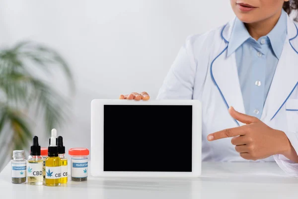 Vista recortada del médico afroamericano señalando con el dedo a la tableta digital con pantalla en blanco cerca de botellas con cannabis medicinal - foto de stock