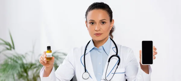 Африканский американский врач держит бутылку с CBD и смартфон с пустым экраном, баннер — стоковое фото