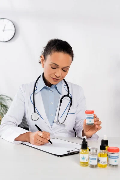 Brunette african american doctor with stethoscope holding bottle with medical cannabis lettering and writing on clipboard — Stock Photo