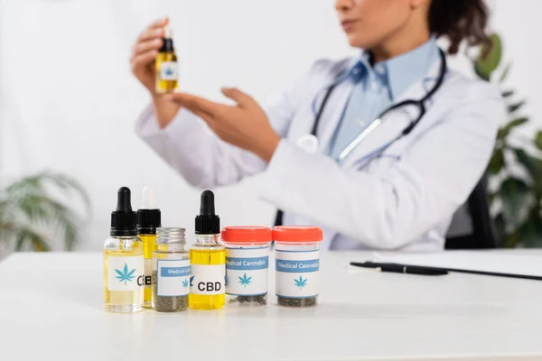 Bottles with cbd and medical cannabis lettering near african american doctor on blurred background — Stock Photo