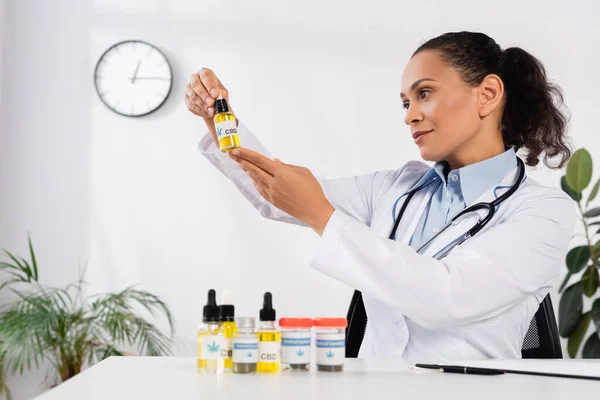 Brunette african american doctor with stethoscope looking at bottle with cbd lettering — Stock Photo