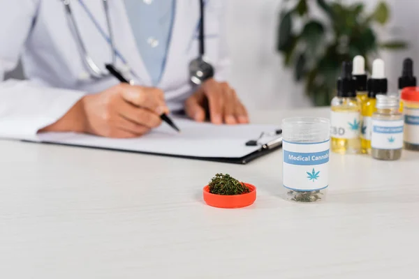 Bottle with medical cannabis lettering and green dried weed near doctor writing prescription on blurred background — Stock Photo