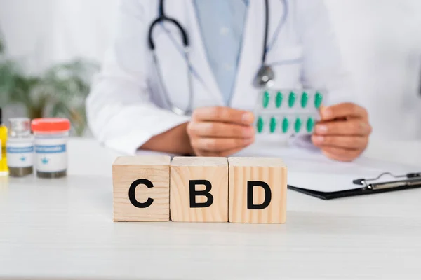 Cubos de madera con letras cbd cerca del médico sosteniendo blíster con cápsulas sobre fondo borroso - foto de stock