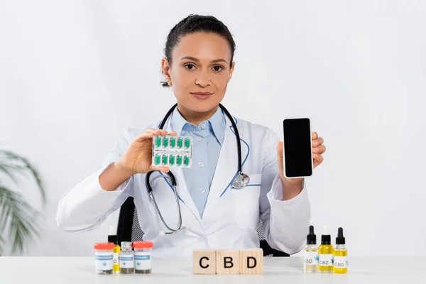 African american doctor with stethoscope holding blister pack with capsules and smartphone near cbd cubes and bottles — Stock Photo