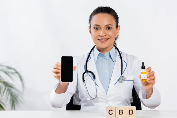 Happy african american doctor holding bottle and smartphone with blank screen near cbd cubes — Stock Photo