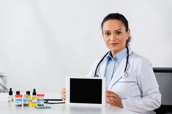 Médico afroamericano sosteniendo tableta digital con pantalla en blanco cerca de la medicación cbd en el escritorio - foto de stock