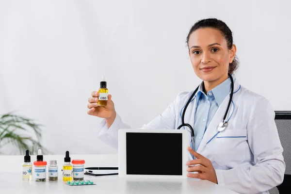 Médico afroamericano feliz celebración tableta digital con pantalla en blanco y botella de cbd cerca de la medicación en el escritorio - foto de stock