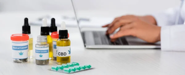 Cropped view of african american doctor typing on laptop near cannabis medication on desk, banner — Stock Photo