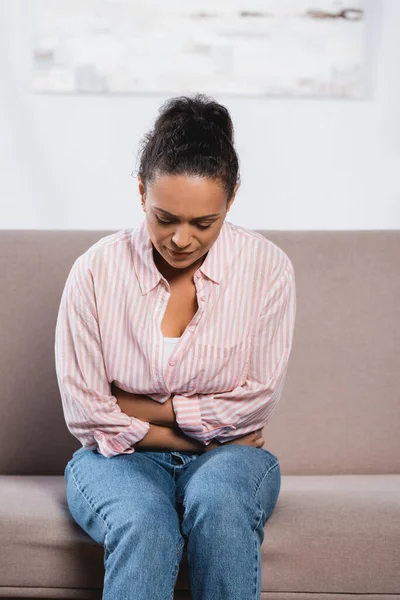 African american woman suffering from abdominal pain while sitting on sofa in living room — Stock Photo