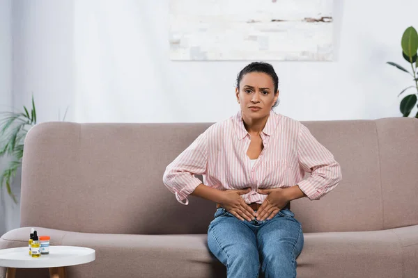 African american woman suffering from abdominal pain while sitting on couch in living room — Stock Photo