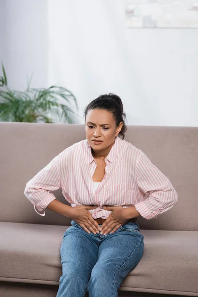 Afro-americana sofrendo de dor abdominal enquanto sentado no sofá — Fotografia de Stock