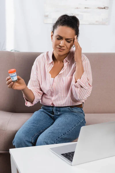 African american freelancer suffering from headache and holding bottle with medical cannabis near laptop — Stock Photo