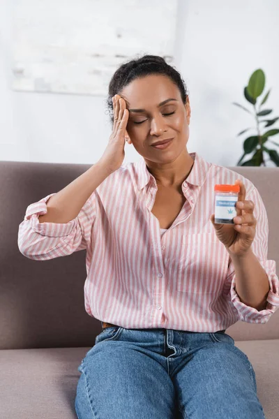 African american woman with closed eyes suffering from migraine and holding bottle with medical cannabis — Stock Photo