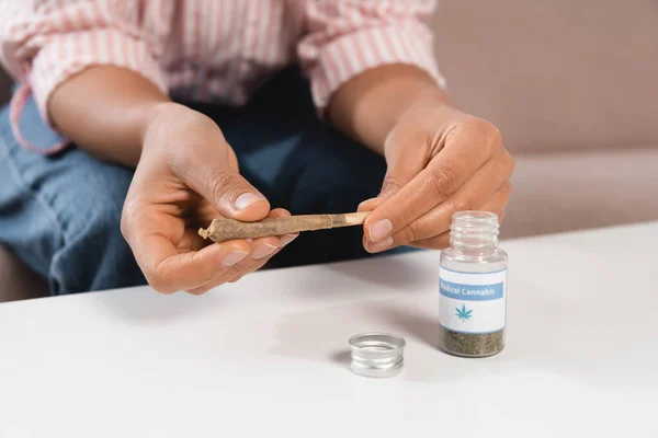 Abgeschnittene Ansicht einer afrikanisch-amerikanischen Frau, die einen Joint in der Nähe einer Flasche mit medizinischem Cannabis-Schriftzug auf dem Tisch hält — Stockfoto