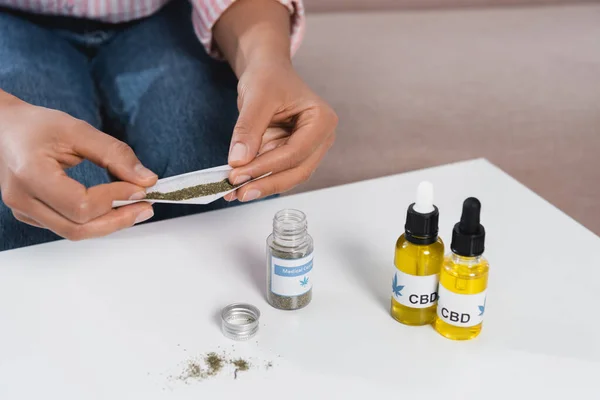 Zugeschnittene Ansicht einer afrikanisch-amerikanischen Frau, die Papier mit getrocknetem medizinischem Cannabis in der Nähe von cbd-Flaschen rollt — Stockfoto