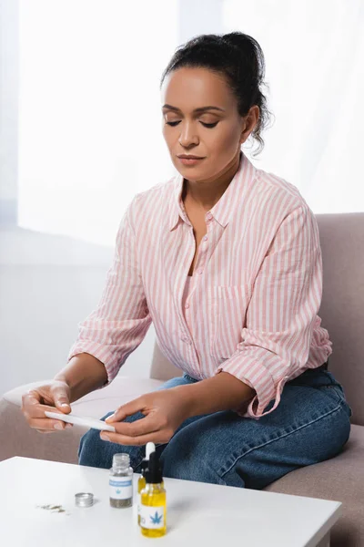 African american woman sitting on couch while rolling paper with dried medical cannabis near cbd bottles — Stock Photo