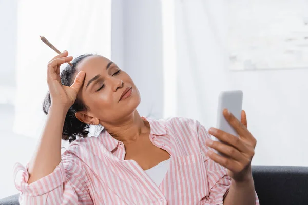 African american woman holding rolled joint with medical cannabis and using smartphone — Stock Photo