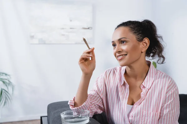 Happy african american woman holding rolled joint with medical cannabis near ashtray — Stock Photo