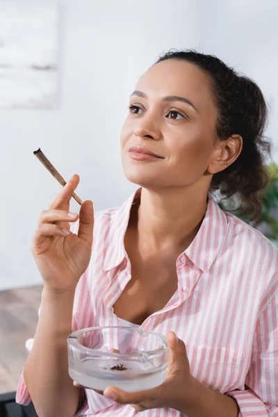 Pleased african american woman holding joint with medical cannabis and ashtray — Stock Photo