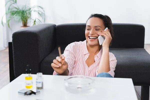 Mulher americana africana feliz falando no smartphone enquanto fumava conjunta perto de garrafas com cannabis medicinal em primeiro plano — Fotografia de Stock
