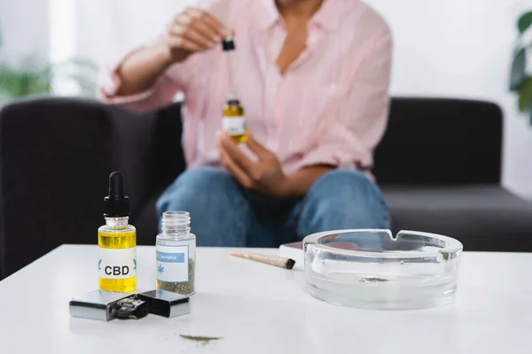 Bottles with cbd, medical cannabis, joint and ashtray on table near african american woman on blurred background — Stock Photo