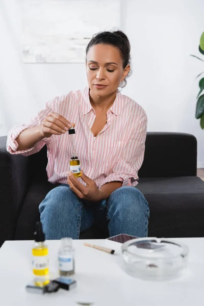African american woman holding bottle with cbd lettering and pipette in living room with blurred foreground — Stock Photo