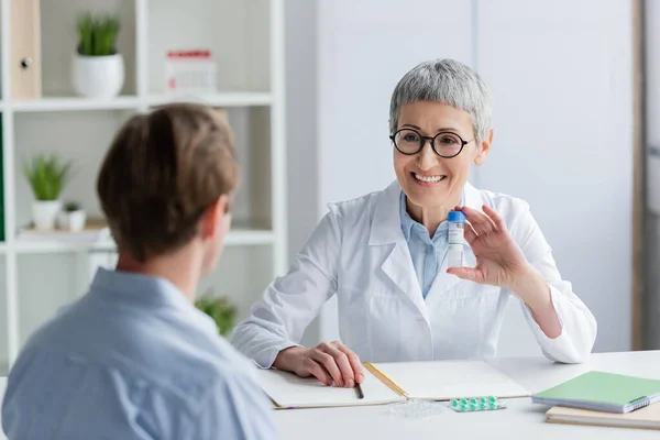 Médico sonriente sosteniendo vacuna cerca de cuadernos, pastillas y paciente en primer plano borroso - foto de stock