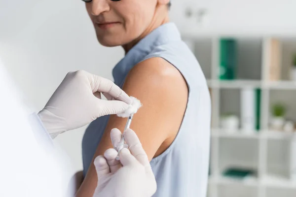 Vista recortada del médico con guantes de látex que sostienen algodón y jeringa cerca de la mujer sobre fondo borroso - foto de stock