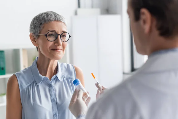 Mujer sonriente que mira al médico con jeringa y vacuna en primer plano borroso - foto de stock