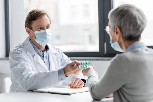Врач в медицинской маске дает пациенту таблетки на размытом переднем плане в клинике — стоковое фото