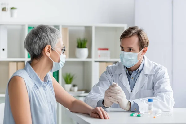 Médico con máscara médica sentado cerca de paciente maduro y vacuna con pastillas durante la consulta - foto de stock