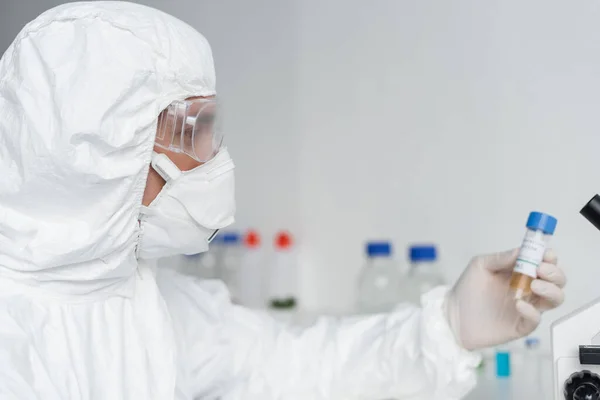 Вчений у захисному костюмі та окулярах тримає вакцину на розмитому фоні — стокове фото