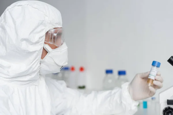 Científico en traje de protección y gafas que sostiene la vacuna sobre fondo borroso - foto de stock
