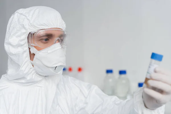 Scientist in protective suit and mask holding vaccine on blurred foreground — Stock Photo