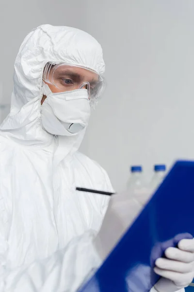Scientist in hazmat suit and medical mask writing on clipboard on blurred foreground — Stock Photo
