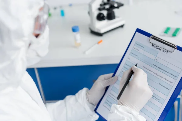 Medical card and pen in hands of scientist in protective uniform on blurred foreground — Stock Photo