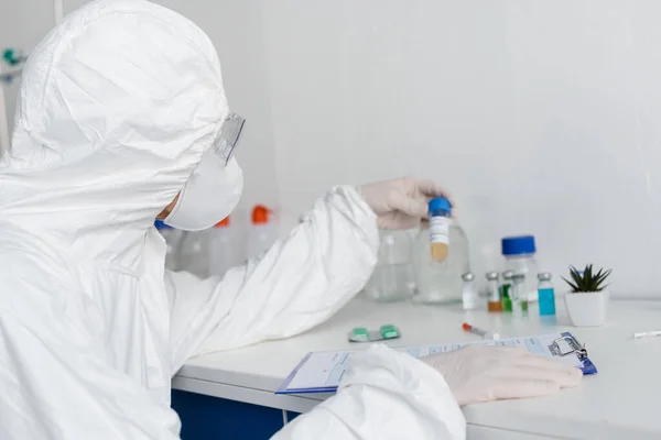 Вчений в захисній формі і латексні рукавички, що тримають вакцину біля буфера обміну на розмитому фоні — стокове фото