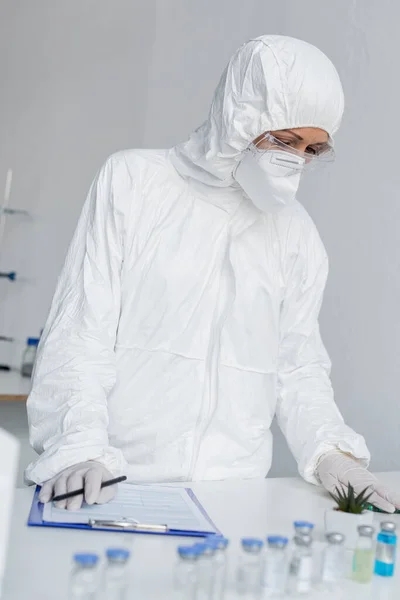 Scientist in hazmat suit standing near vaccines and clipboard on blurred foreground in laboratory — Stock Photo