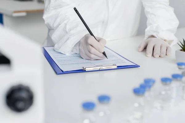 Cropped view of scientist writing on clipboard near vaccines on blurred foreground — Stock Photo