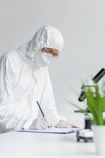 Scientist in protective suit writing on clipboard near microscope on blurred foreground — Stock Photo