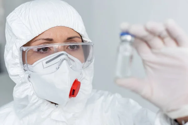 Вчений у захисній масці та окулярах тримає вакцину на розмитому фоні — стокове фото