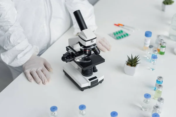 Обрізаний вид вченого в костюмі небезпеки, що стоїть біля мікроскопа і вакцин на столі — стокове фото
