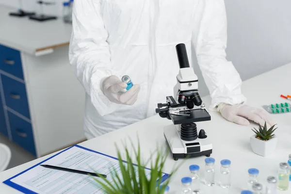 Cropped view of scientist in hazmat suit holding vaccine near microscope and clipboard — Stock Photo
